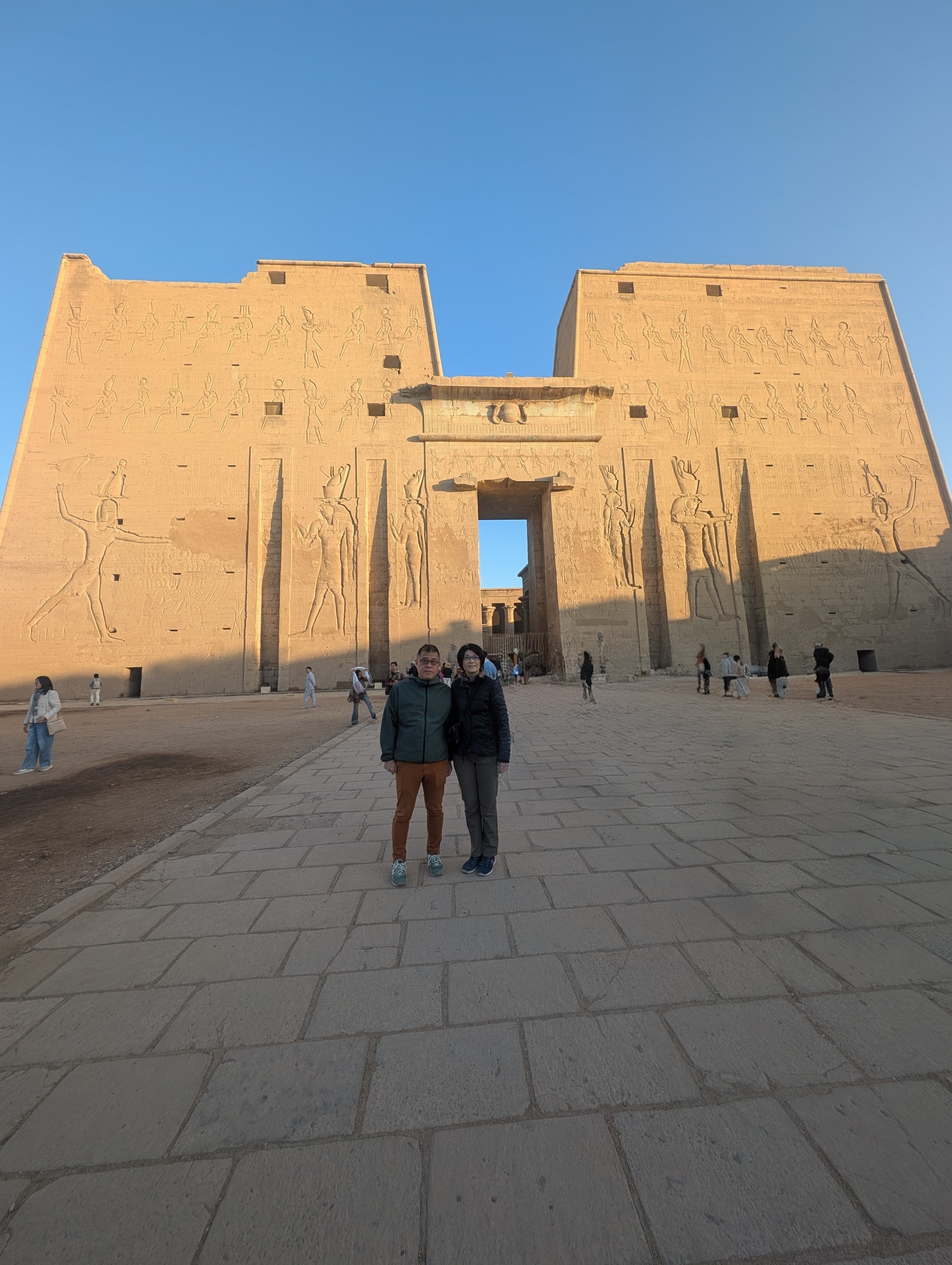 Two visitors posing in front of the Temple of Edfu pylon at golden hour.