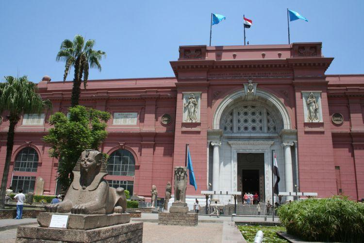 Ancient sphinx statues line the entrance of Cairo's pink-painted Egyptian Museum under clear blue sky.