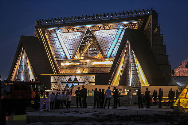 The Grand Egyptian Museum illuminated with golden lights at dusk, crowds gathered outside