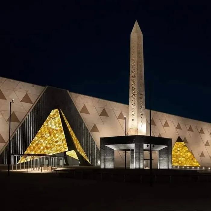 NMEC exterior lit at night with golden pyramid shapes and a tall obelisk centerpiece