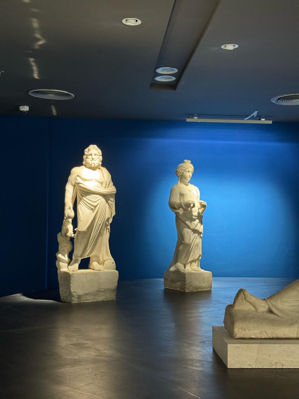 Full-length marble statues of a bearded god and draped female figure under blue gallery light