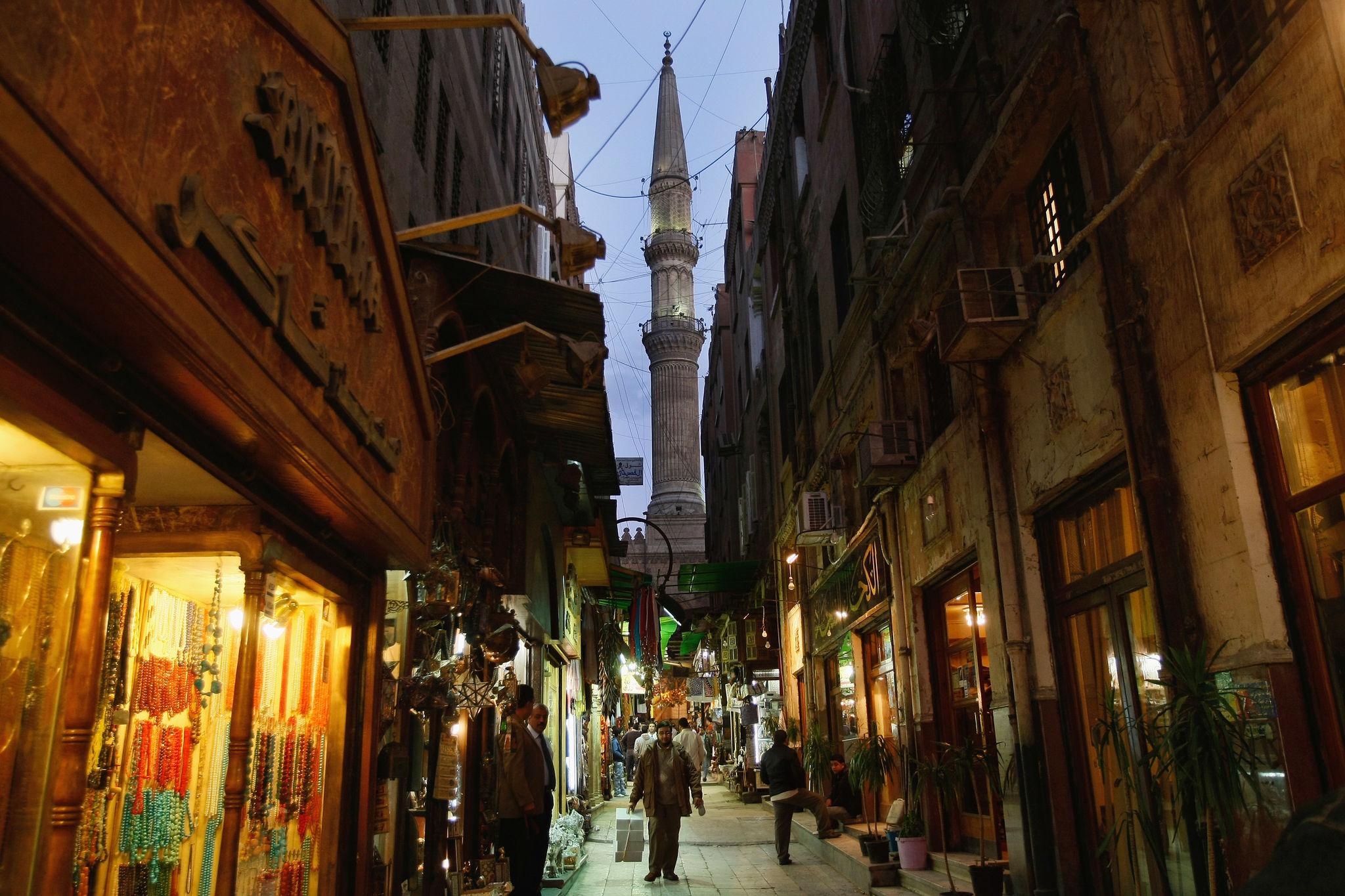 Narrow bazaar alley with glowing jewelry shops and a mosque minaret rising at dusk