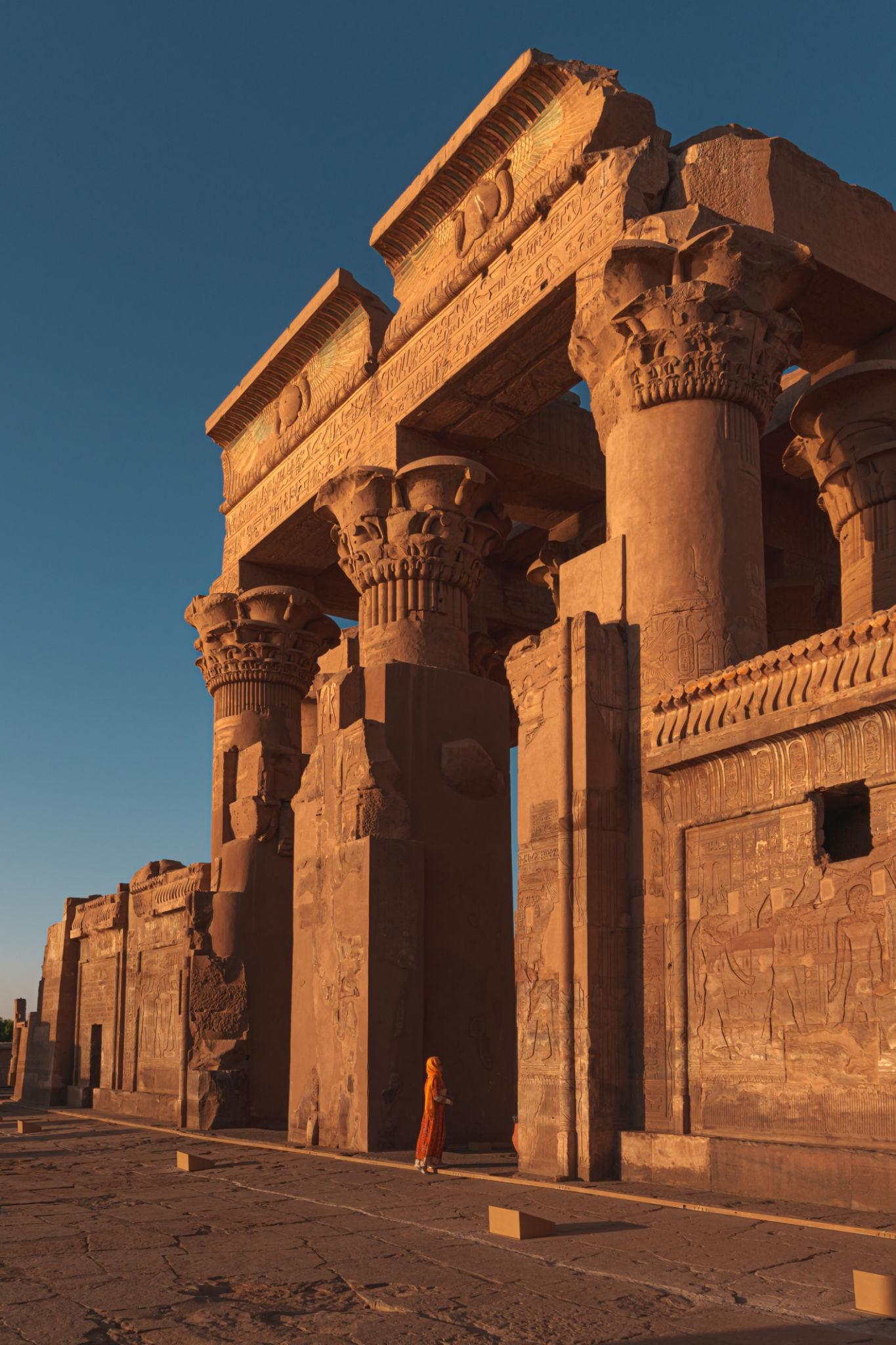 Visitor in red beside the sunlit entrance colonnade of Kom Ombo Temple at sunset.