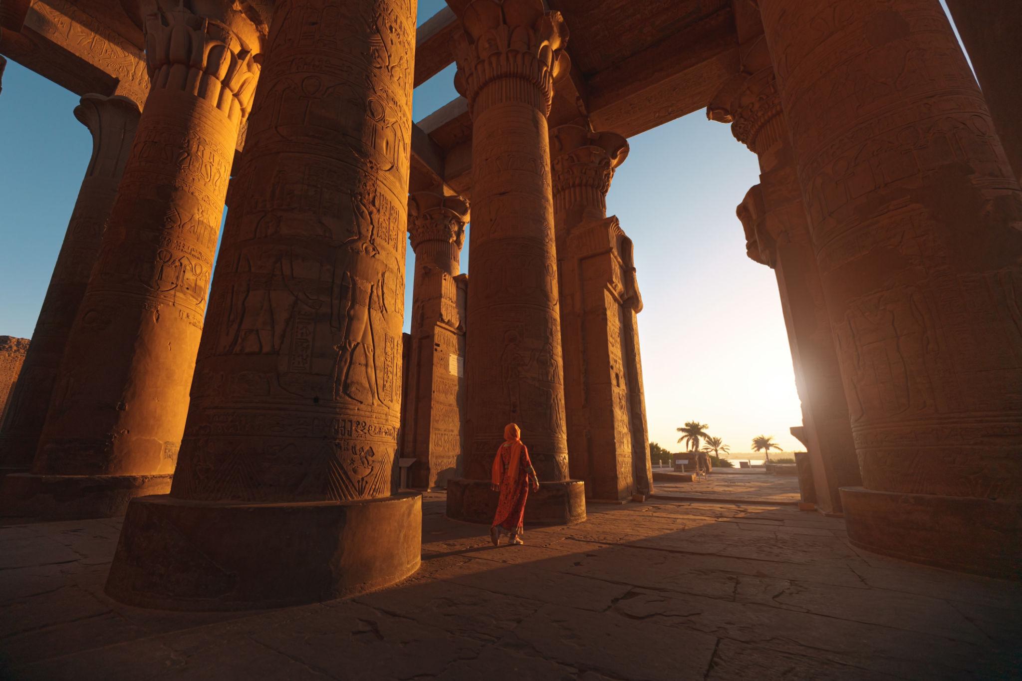 Visitor in red walking among golden-lit temple columns at sunset in Kom Ombo.