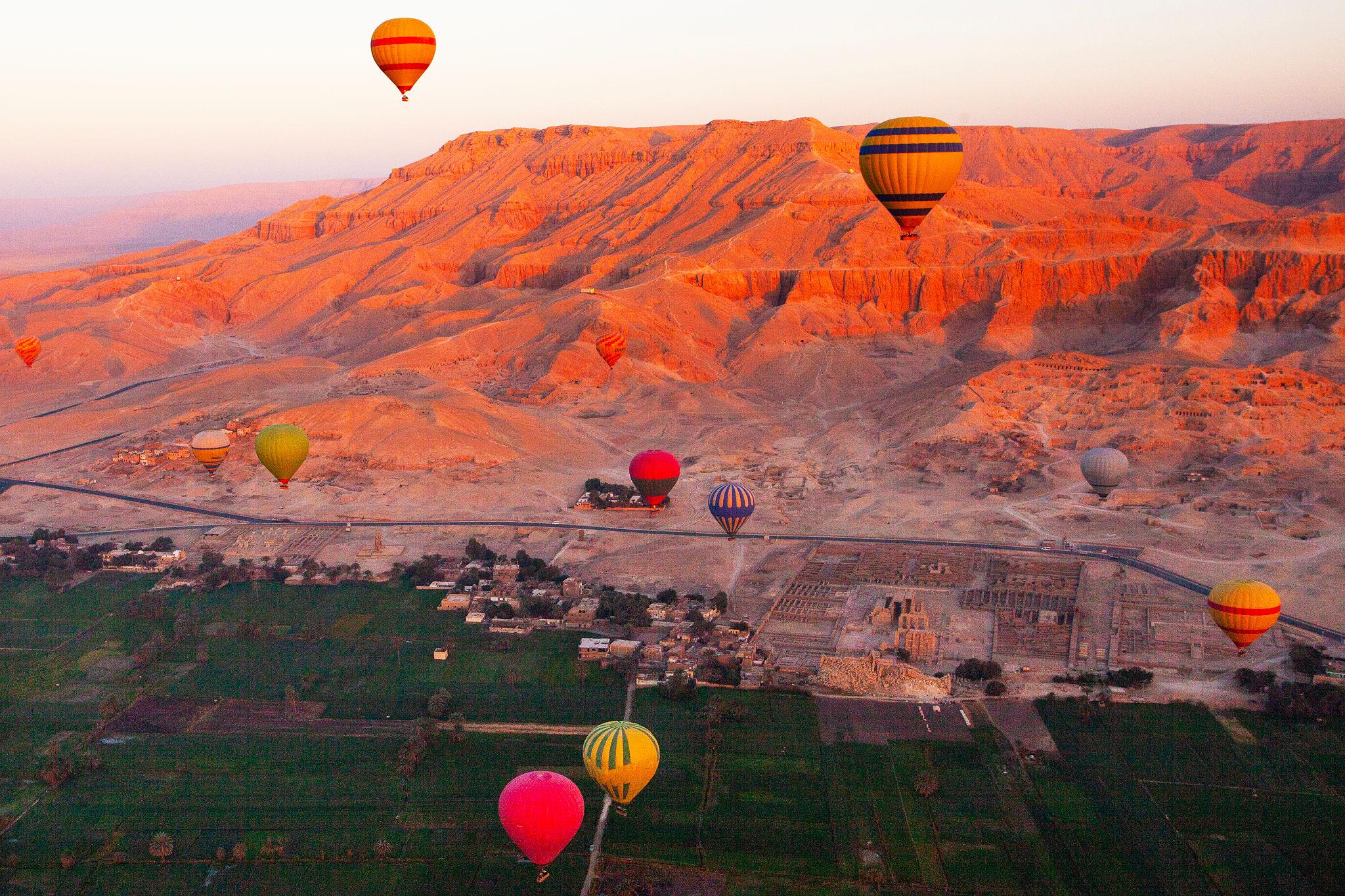 Hot air balloons drifting past red Theban cliffs and the Ramesseum temple ruins at sunrise