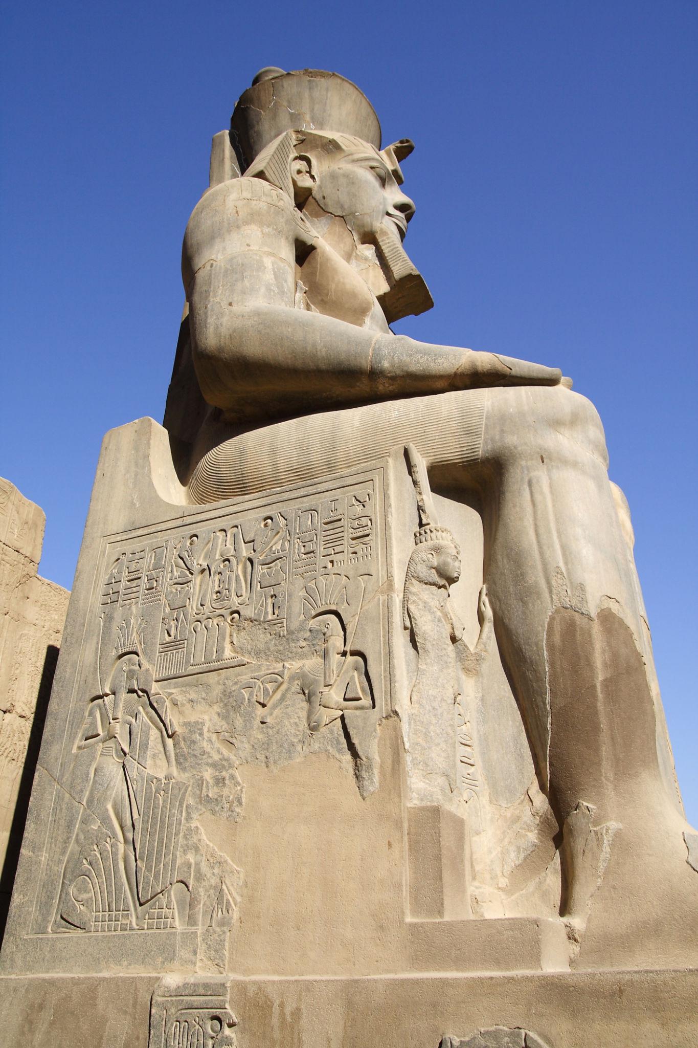 Ramses II seated colossus with carved throne reliefs against blue sky.