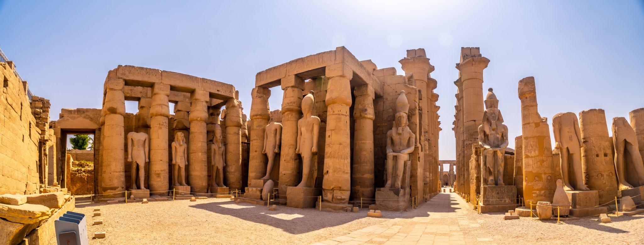 Wide panorama of Luxor Temple courtyard with statues and columns in sunlight.