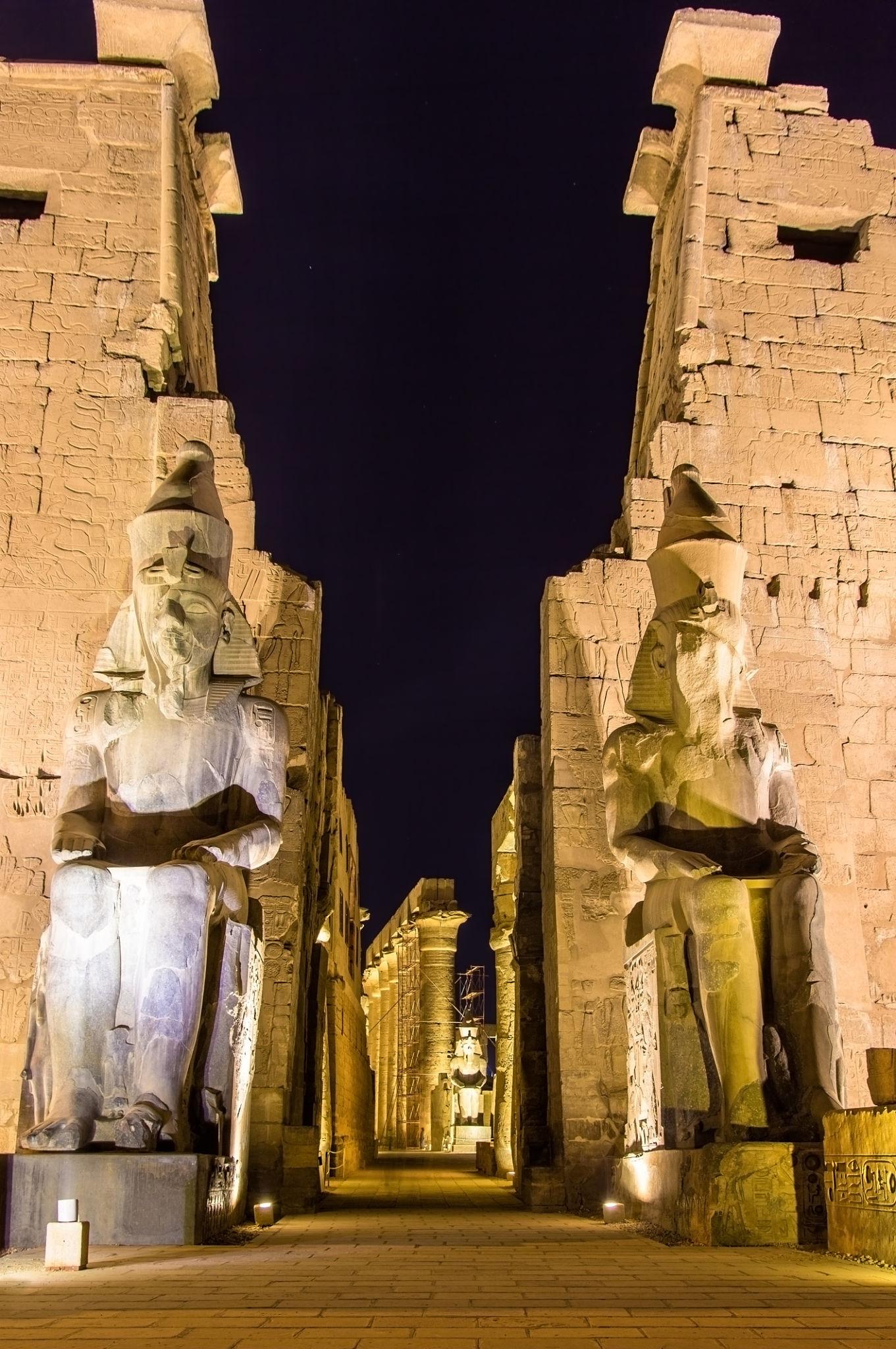 Seated colossi framing the illuminated entry gateway of Luxor Temple.