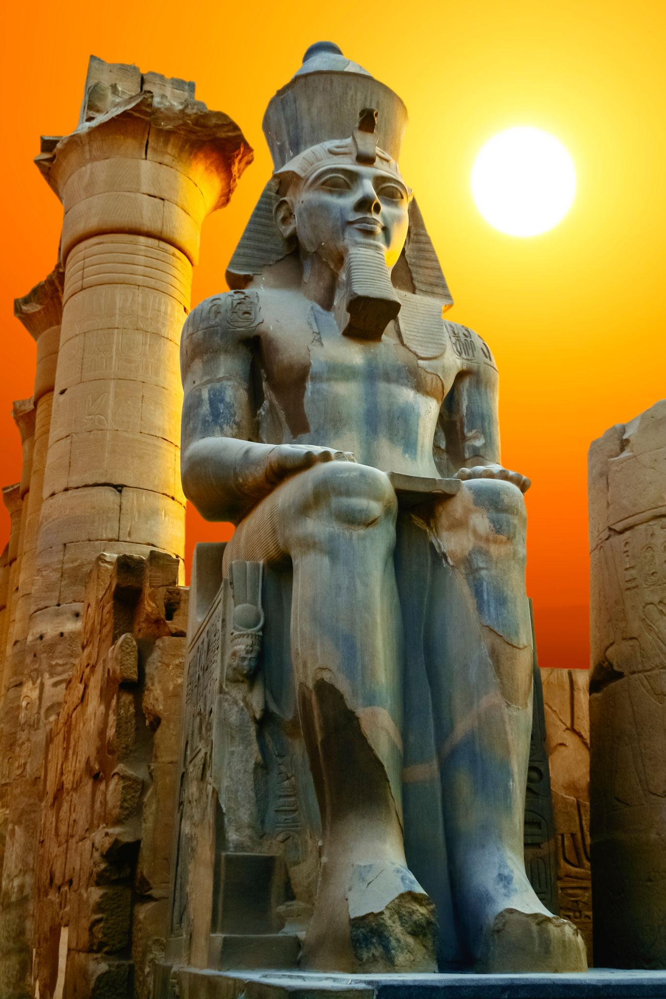Seated Ramses II colossus silhouetted against a blazing orange sun.