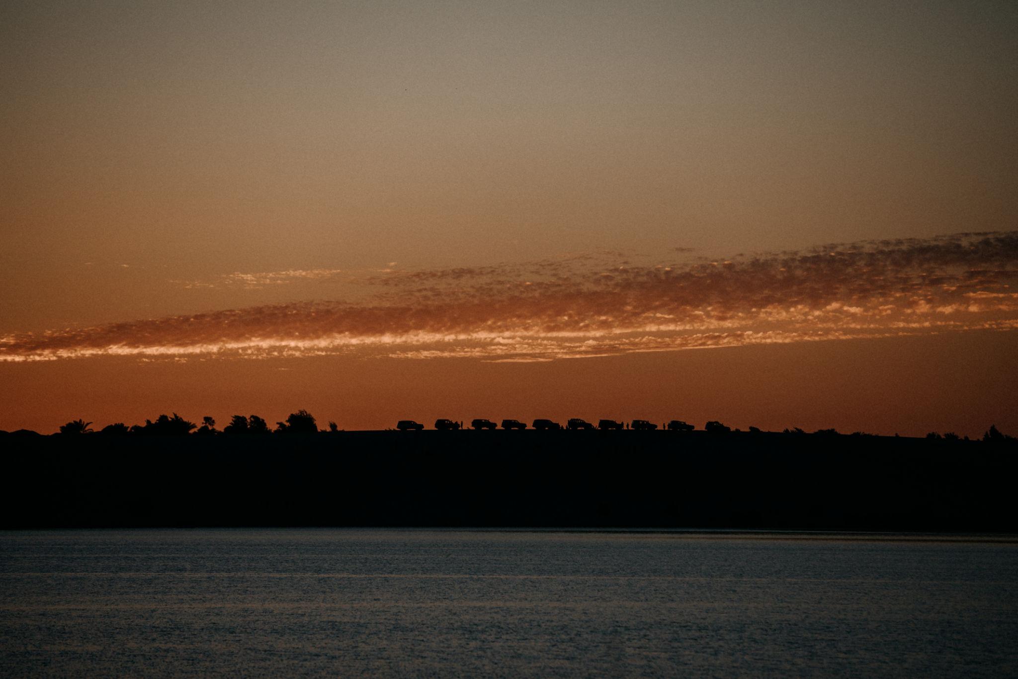 Silhouetted treeline along a calm lake under an orange dusk sky