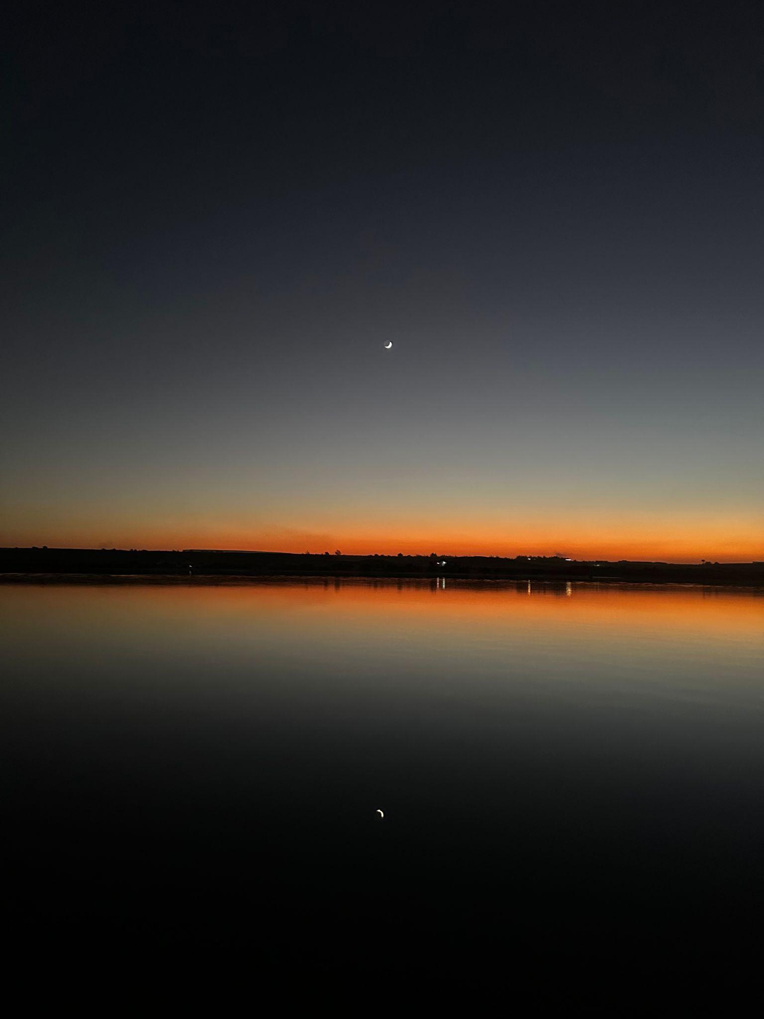 Crescent moon over a still desert lake glowing with orange sunset light
