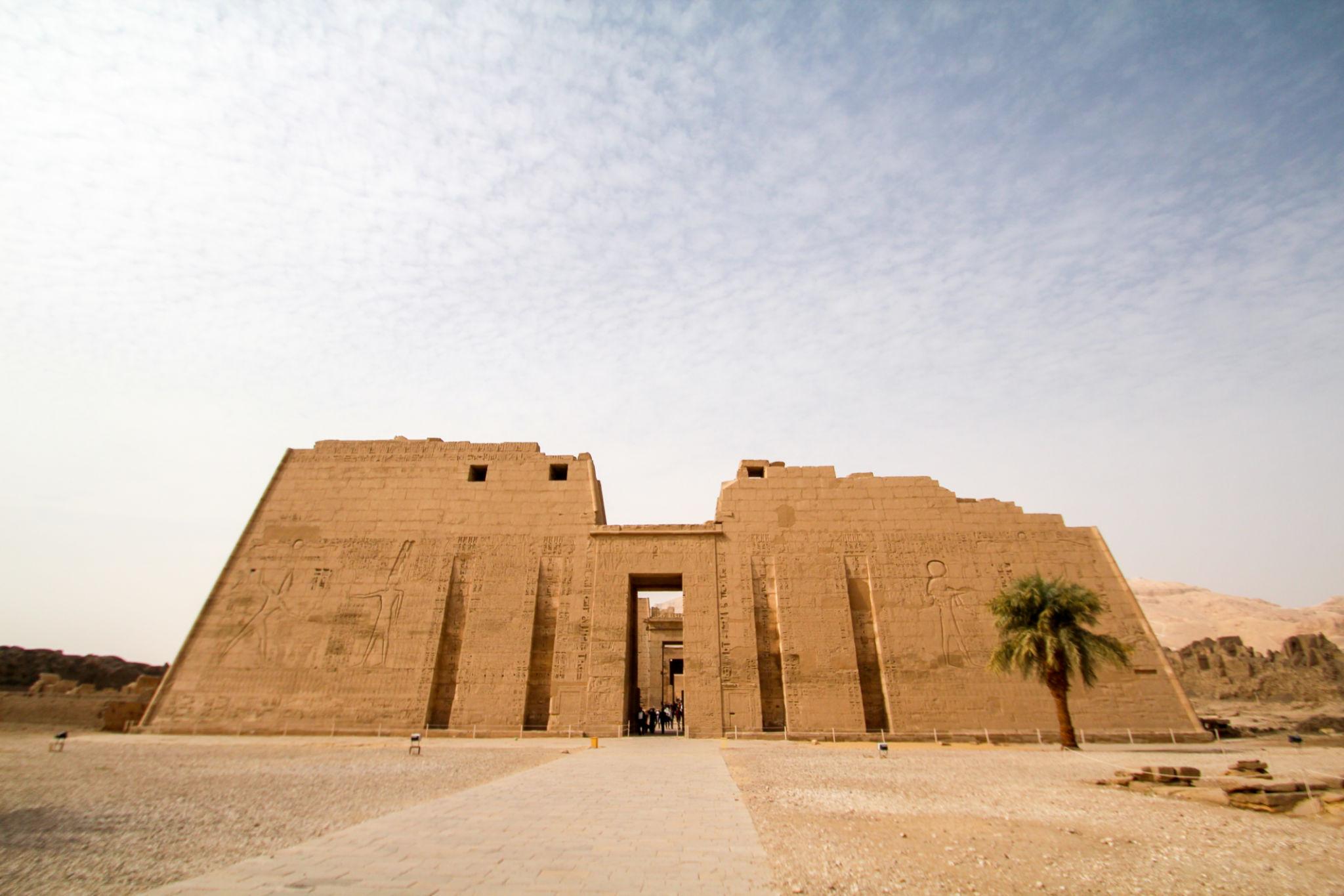 Wide panoramic view of Medinet Habu's first pylon with palm tree and desert sky.