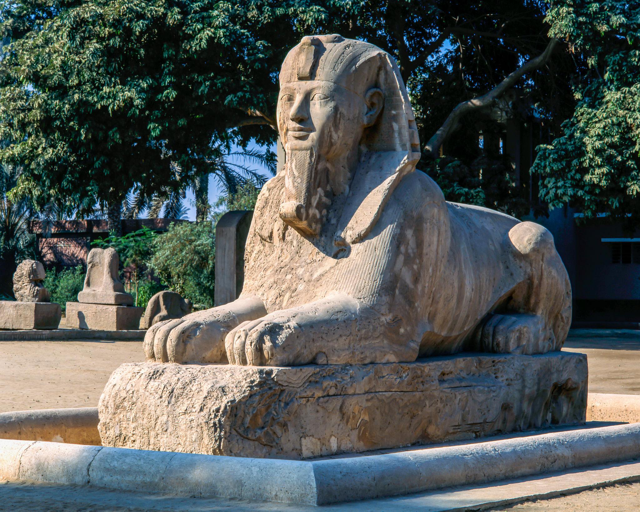 Ancient Egyptian alabaster sphinx statue reclining on a stone base surrounded by trees