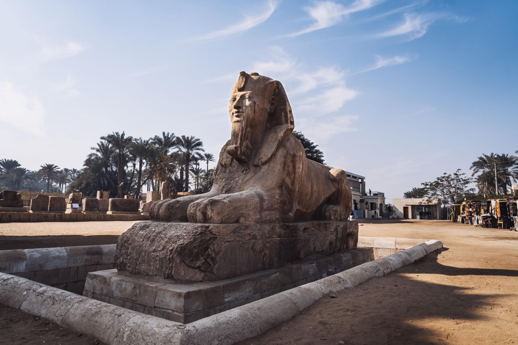 Ancient Egyptian alabaster sphinx statue on a stone plinth surrounded by sandy ground and palm trees