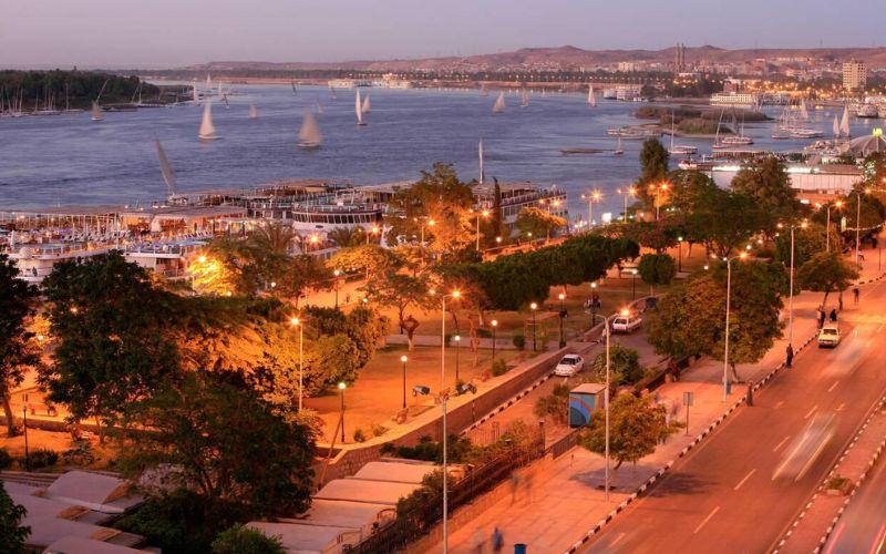Aswan corniche at sunset with feluccas sailing and cruise boats lining the Nile.