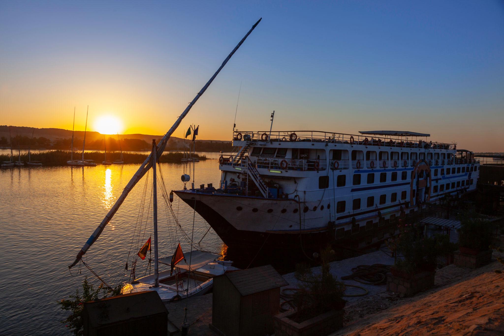 Nile cruise ship docked beside a felucca at sunset over the Aswan riverbank.