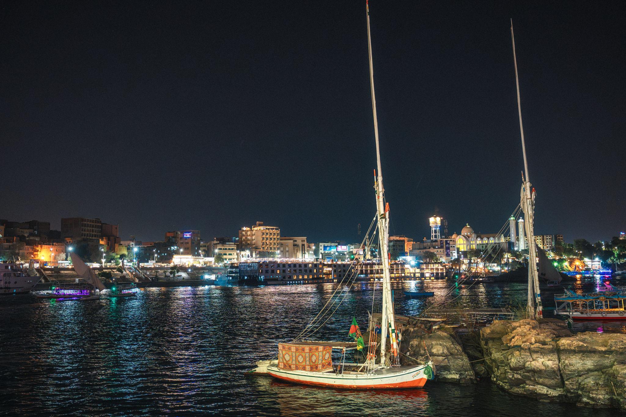 Felucca anchored on the Nile at night with Aswan city lights reflecting on the water.