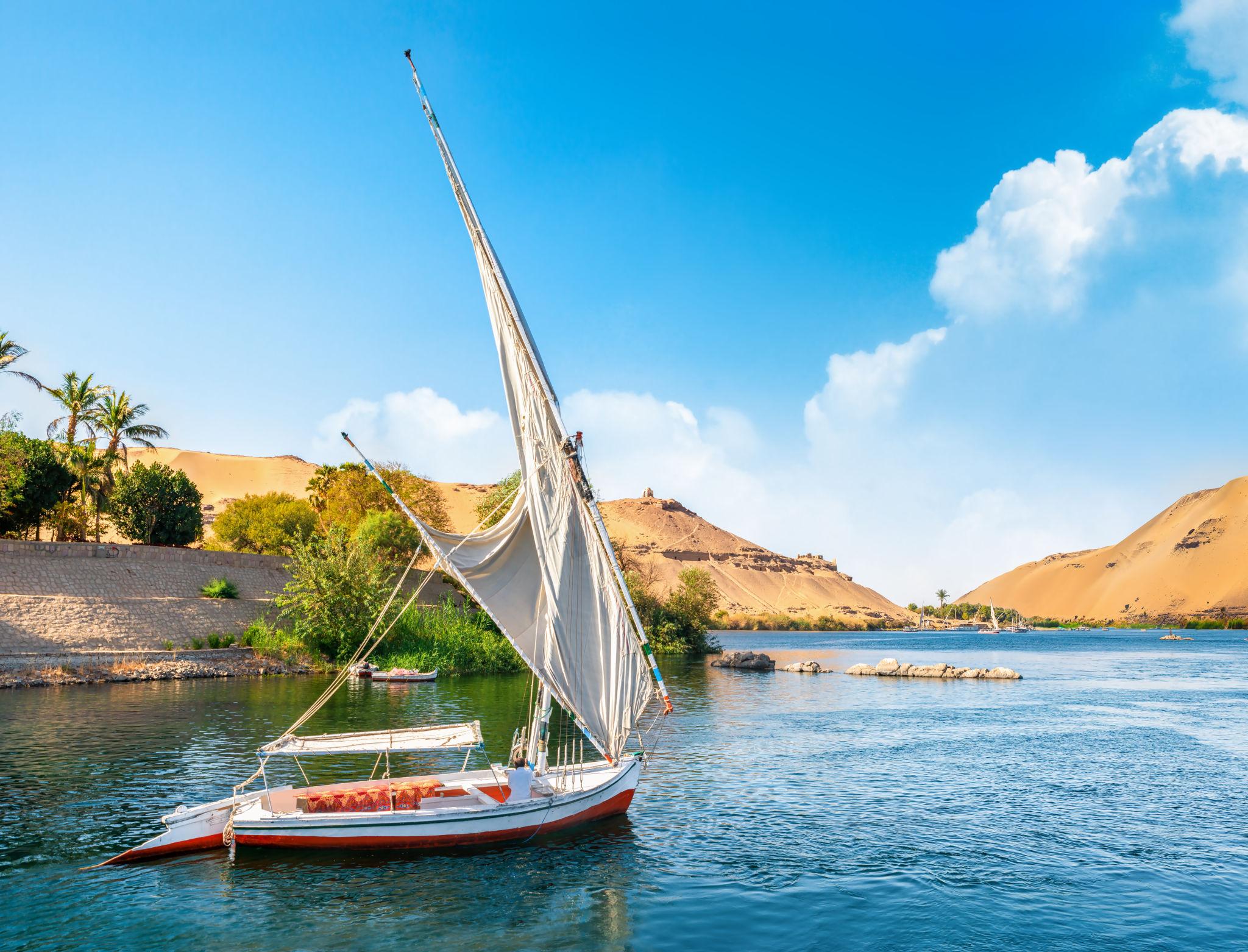 White-sailed felucca gliding past golden sand dunes on the Nile at Aswan.