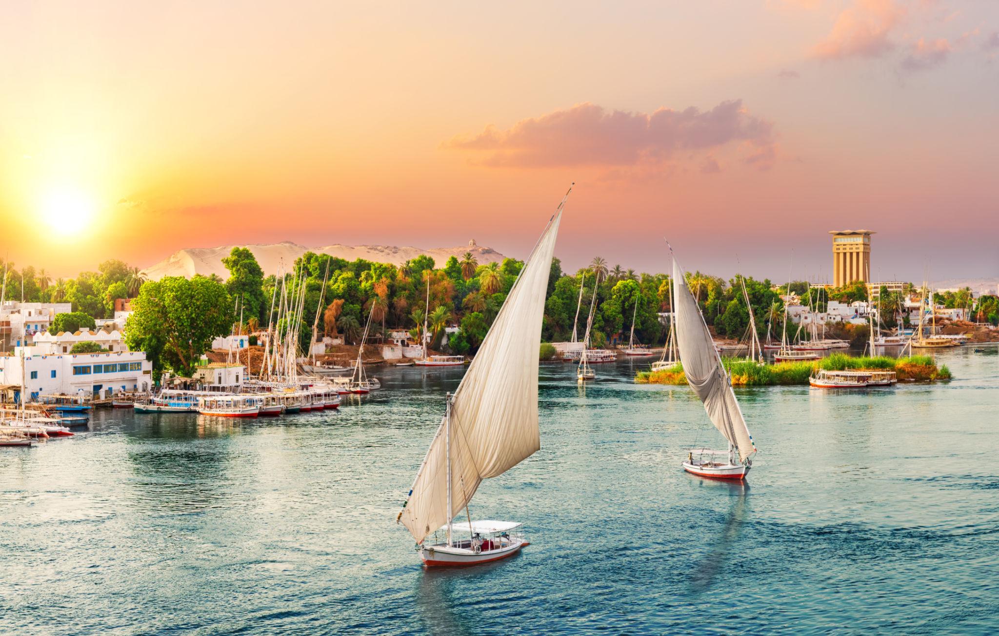 Two feluccas sailing past Aswan riverside buildings under a pastel sunset sky.