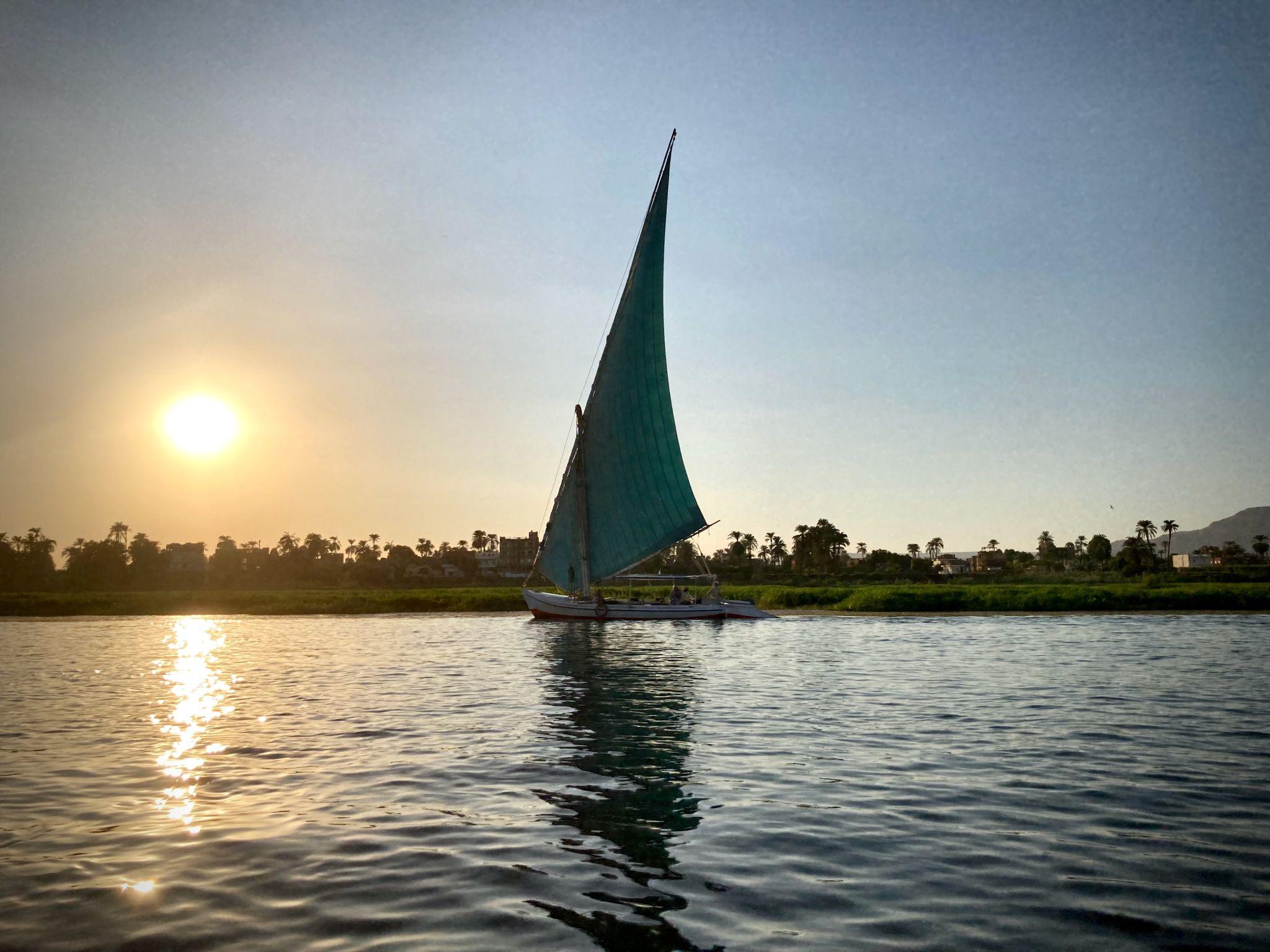 Felucca with teal sail glides past Luxor shore during golden-hour sunset