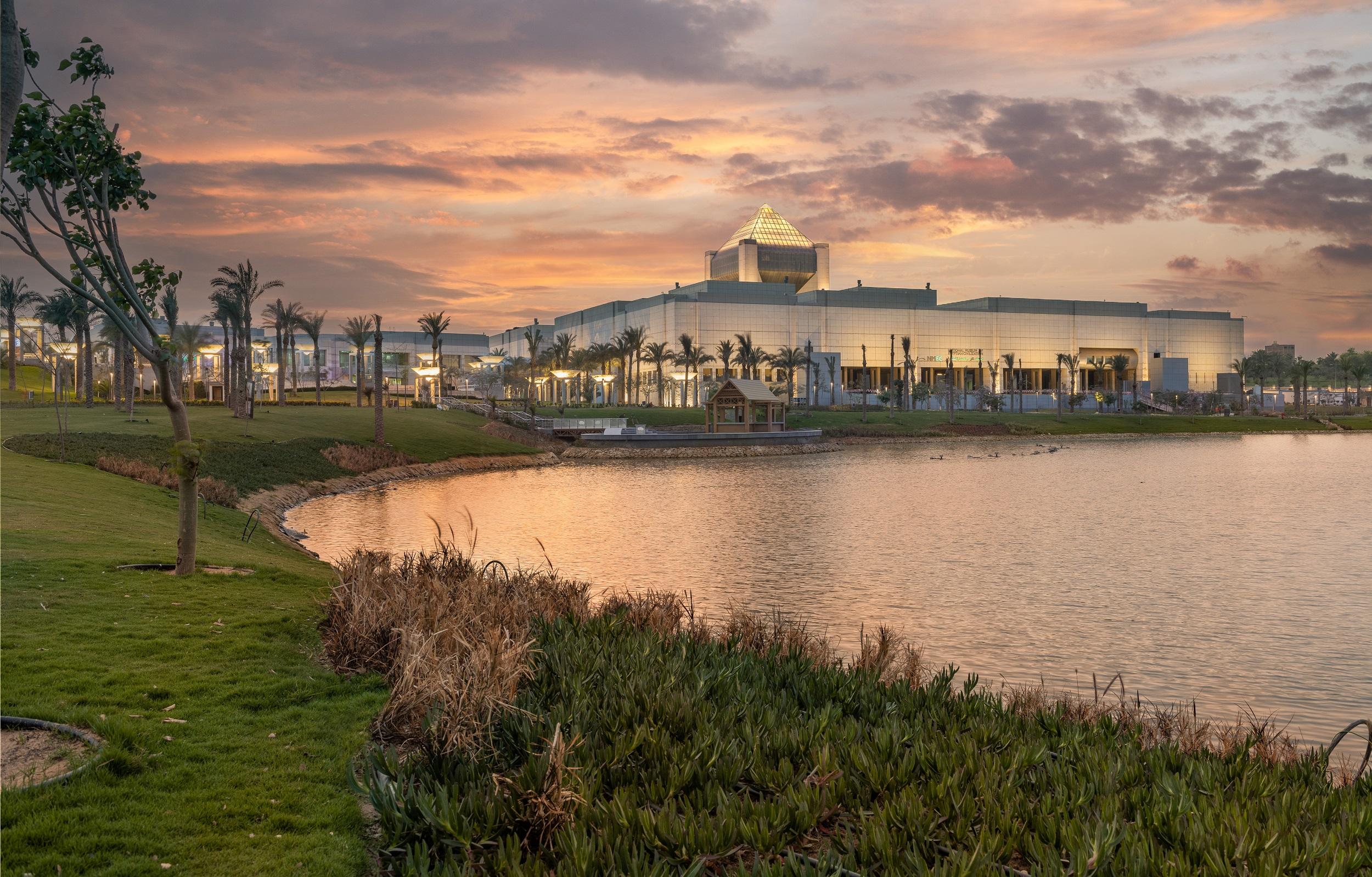 NMEC glass building with golden pyramid roof reflected in a calm lake at sunset