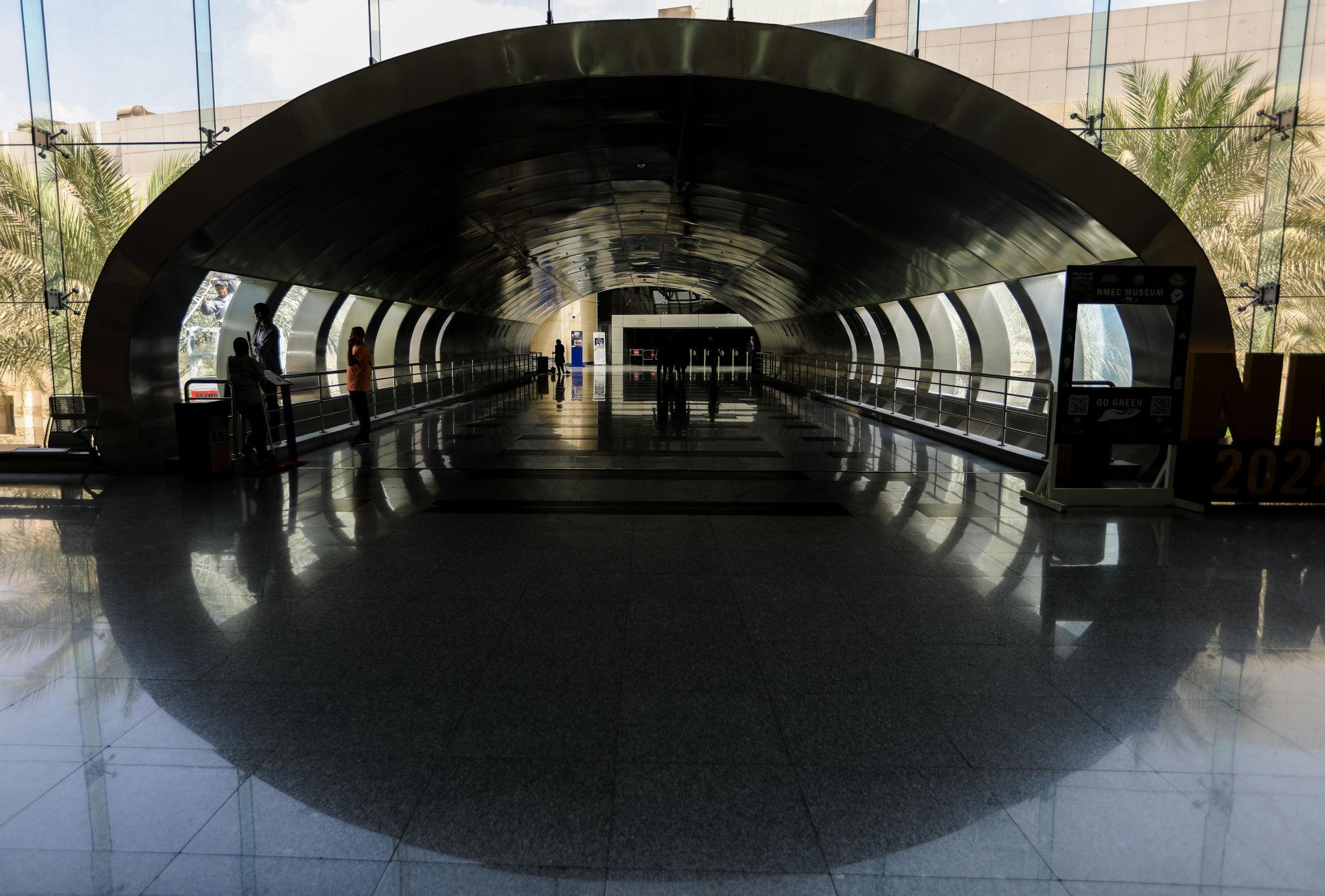 Visitors walk through a dramatic oval steel arch tunnel at the NMEC entrance with reflective floors