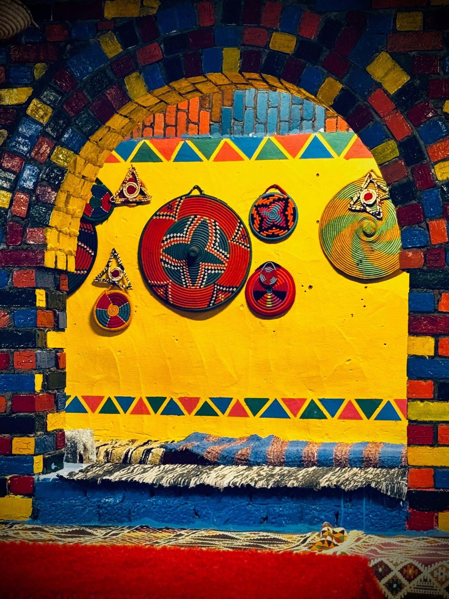 Vibrant woven baskets and geometric crafts displayed on a yellow wall inside a colorful brick arch