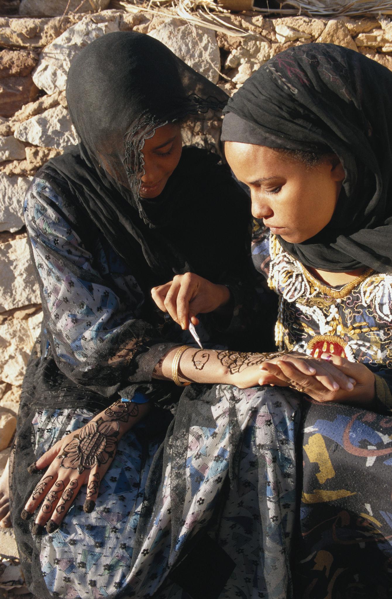 Two women in headscarves, one applying intricate henna designs to the other's hand.