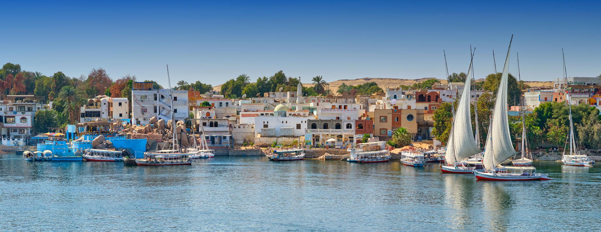 Colorful felucca sailboats glide past a vibrant Egyptian riverside town on the Nile.