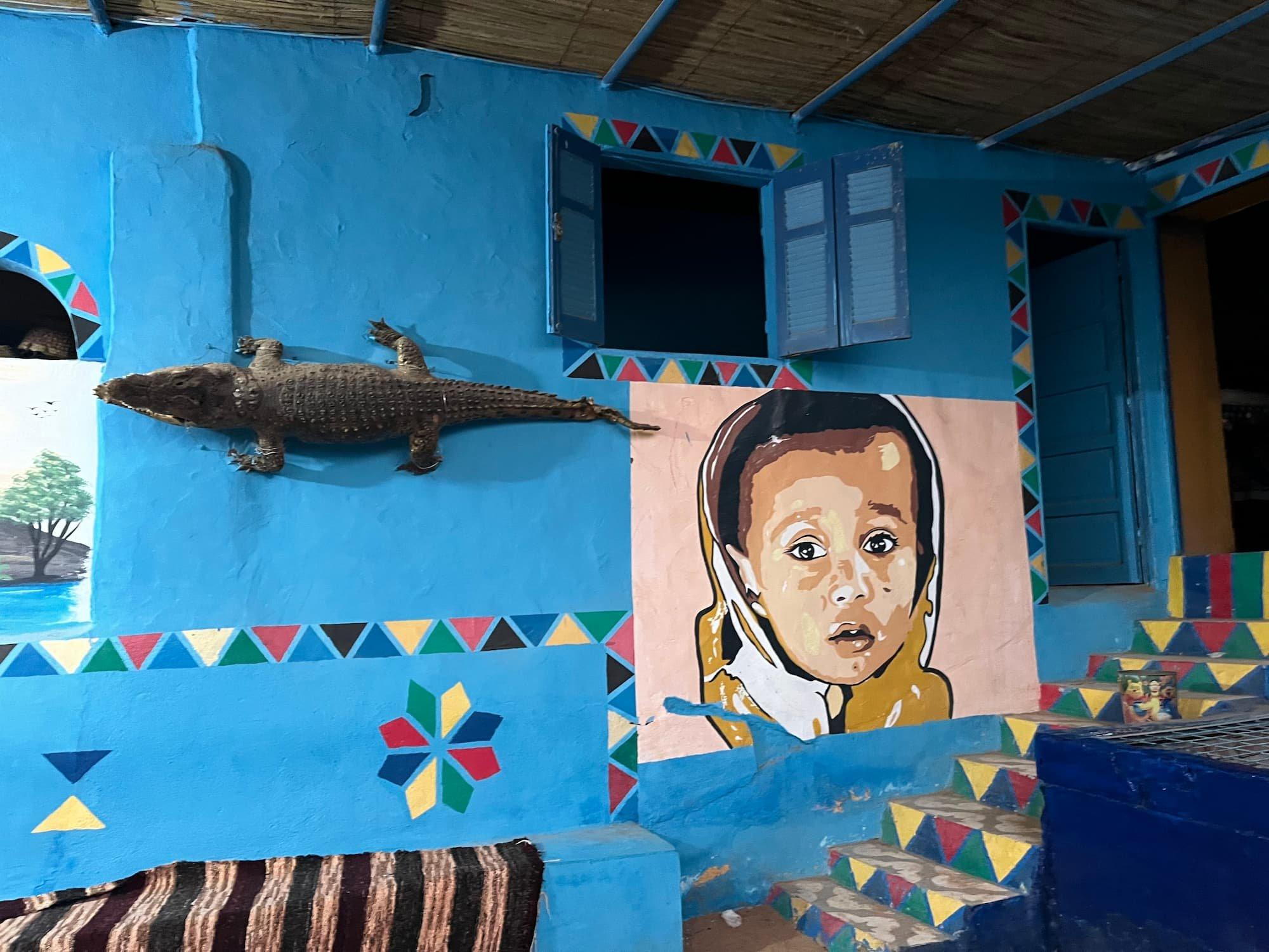 Blue Nubian wall with mounted crocodile and painted child portrait, decorated with colorful geometric patterns