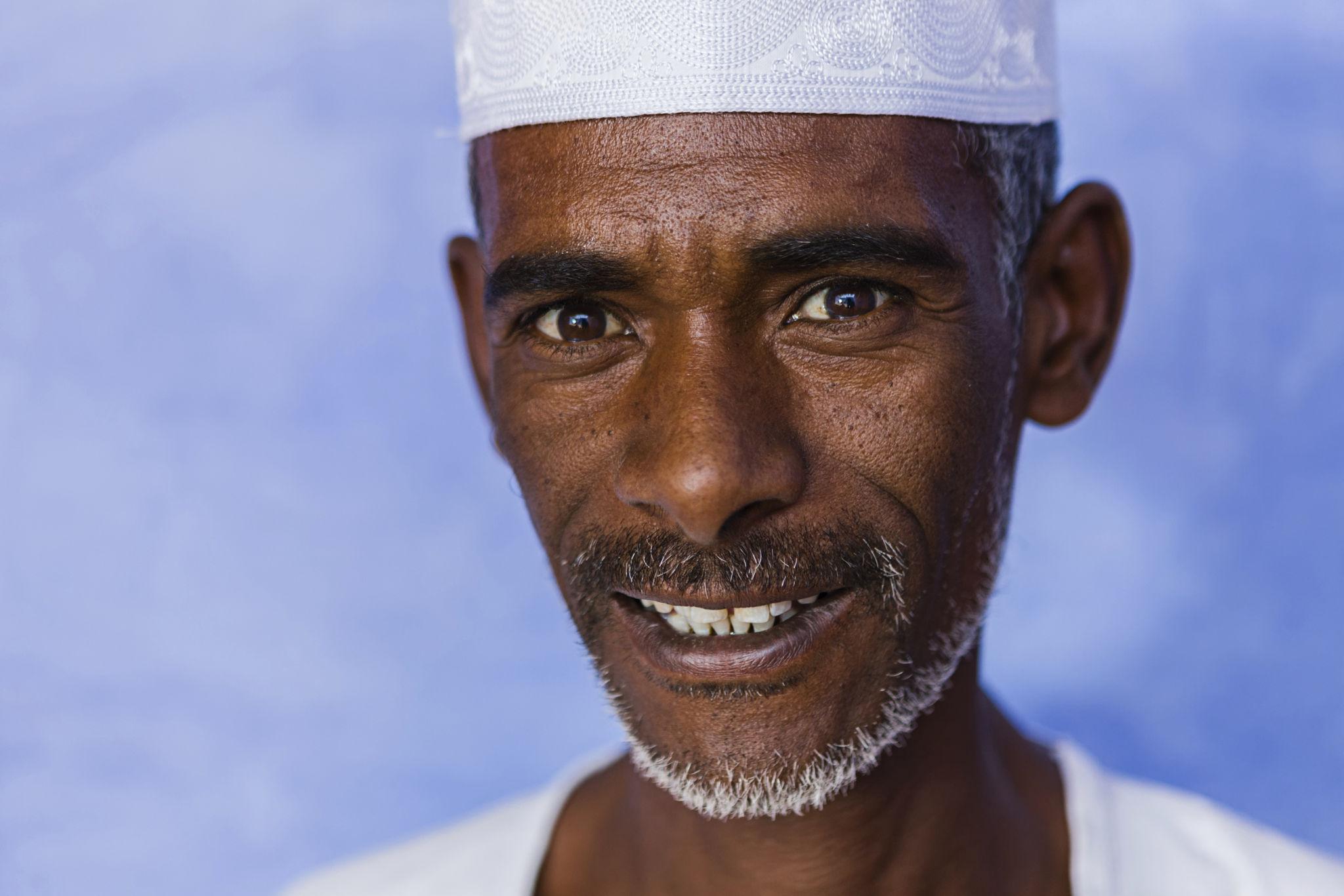 An elderly Egyptian man wearing a white kufi cap smiles warmly against a blue background