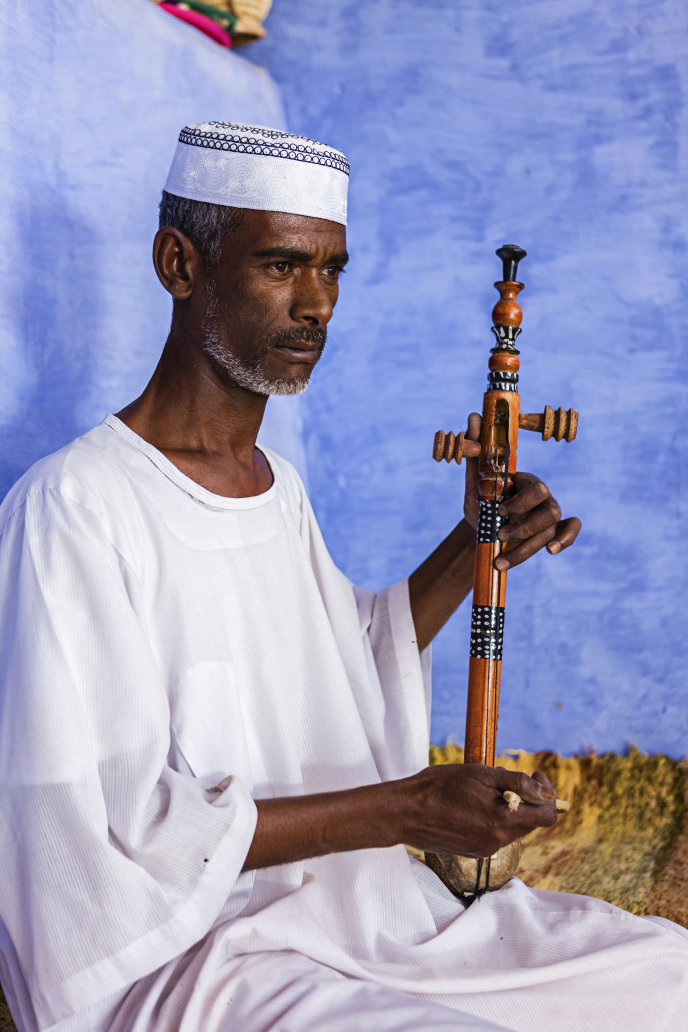 A man in white thobe and kufi cap plays a traditional African string instrument