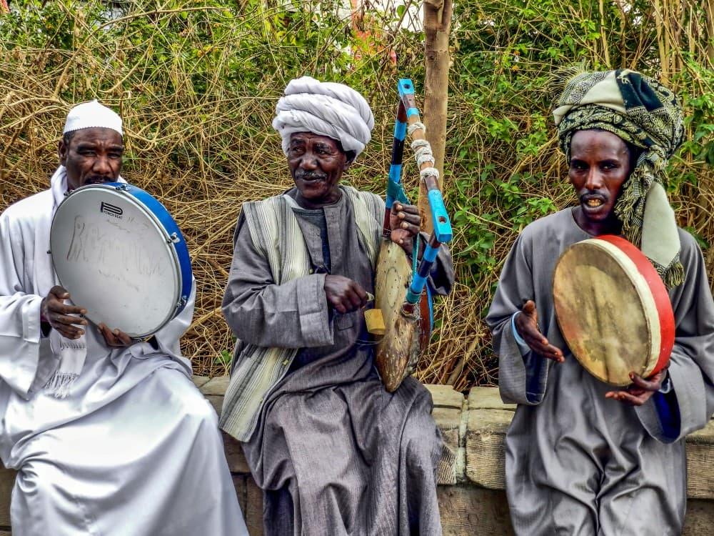 Three Nubian musicians in traditional dress playing drums and a stringed instrument outdoors