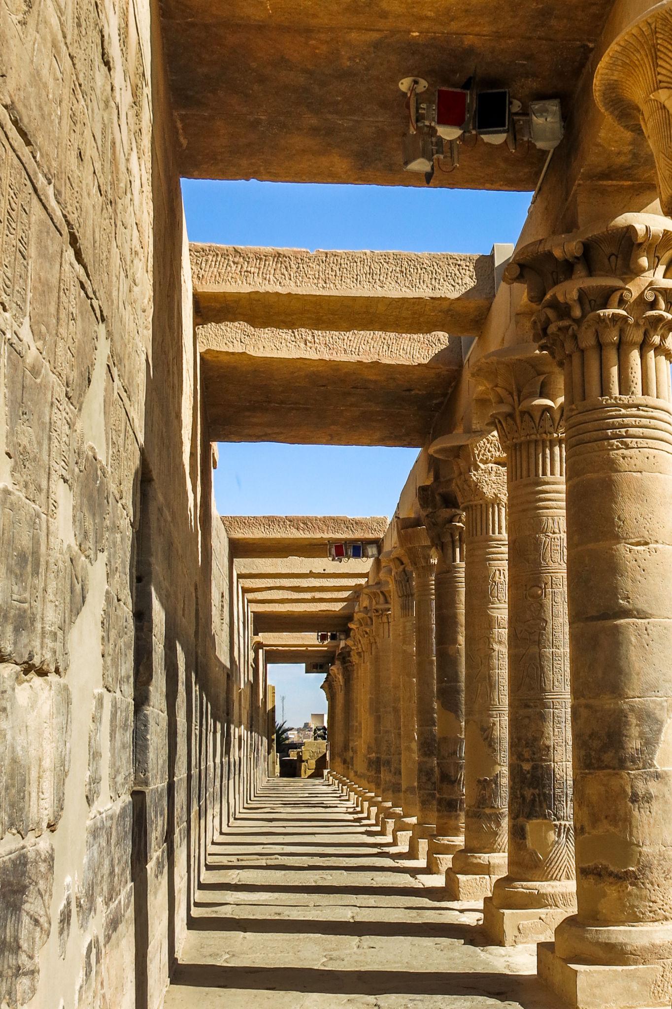 Long colonnaded corridor of Philae Temple with sunlit sandstone columns receding into the distance under a blue sky