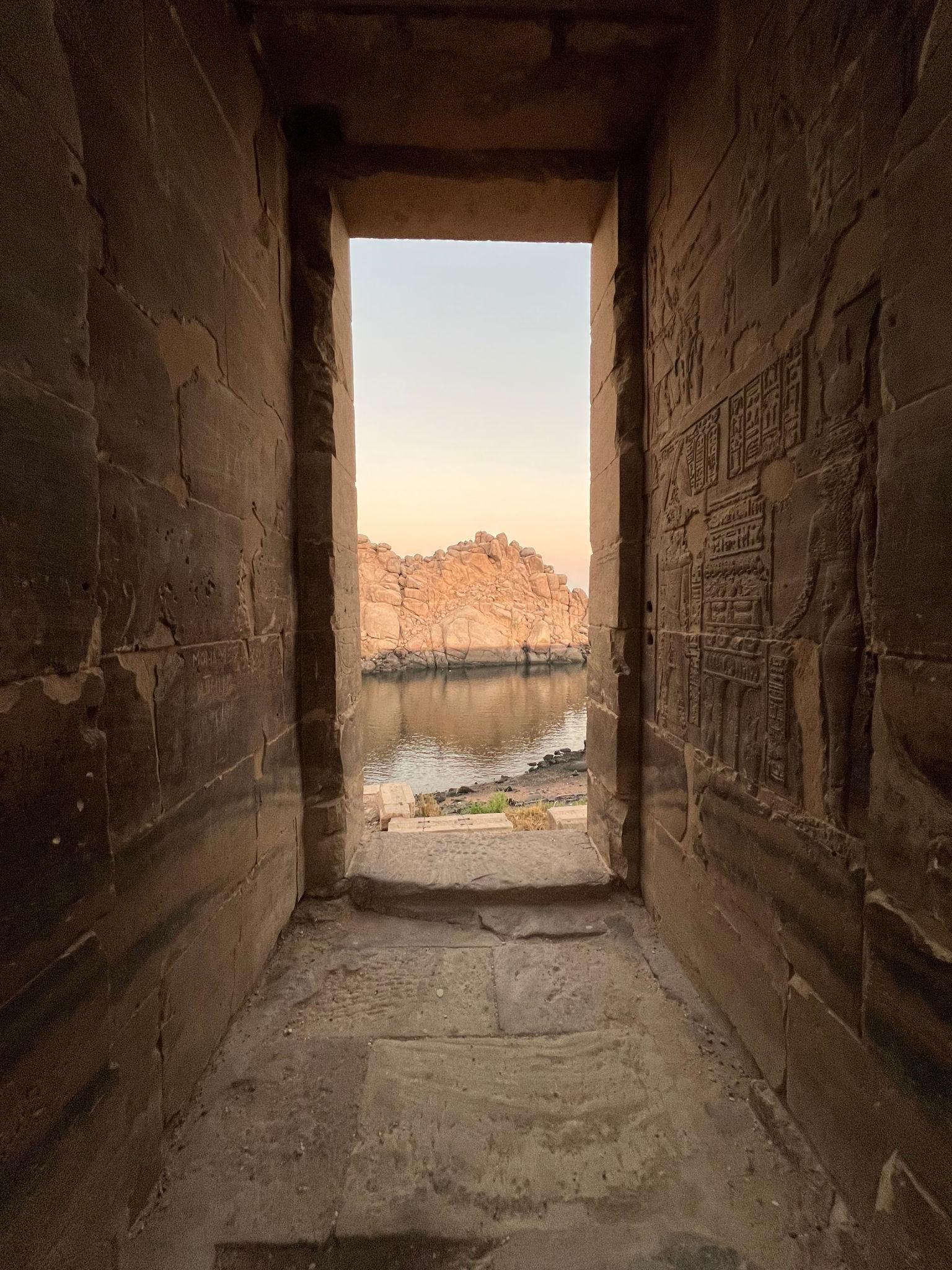 View through a Philae Temple doorway framing the Nile and Aswan cliffs, with hieroglyphic reliefs on the sandstone wall