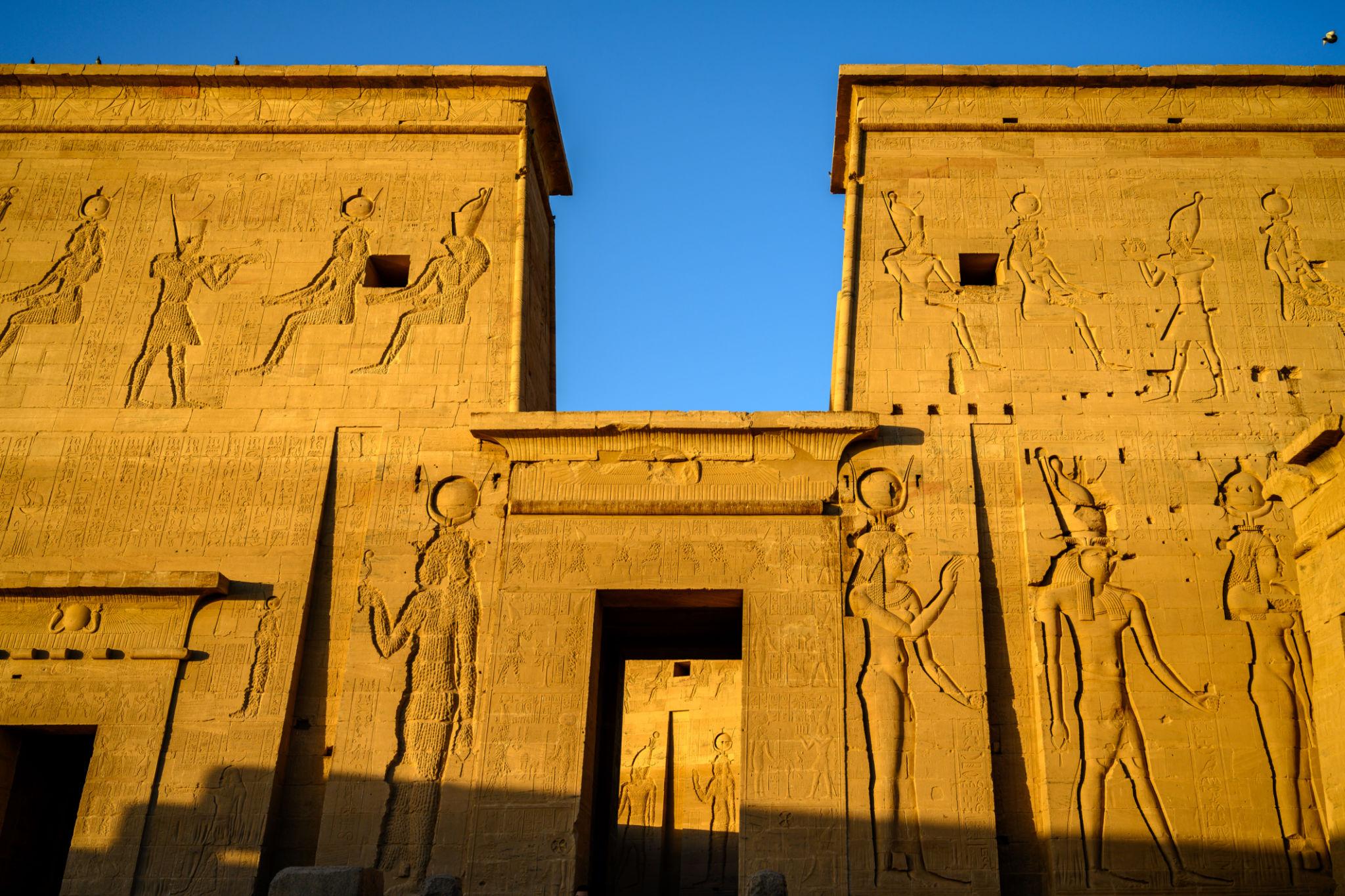 Golden sandstone pylons of Philae Temple covered in carved Egyptian deity reliefs under blue sky