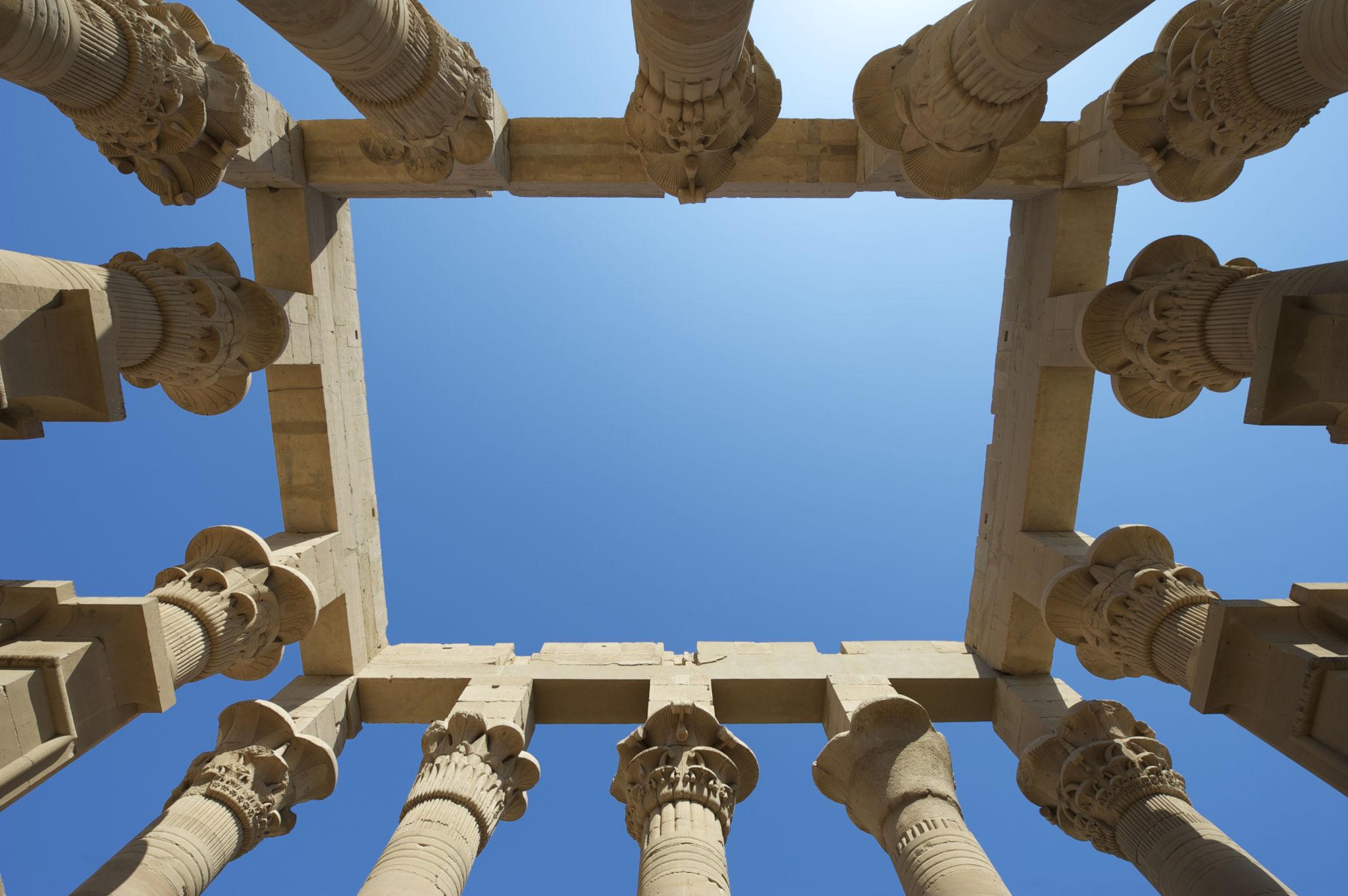 Upward view of ornate ancient Egyptian lotus columns framing a clear blue sky at Philae Temple