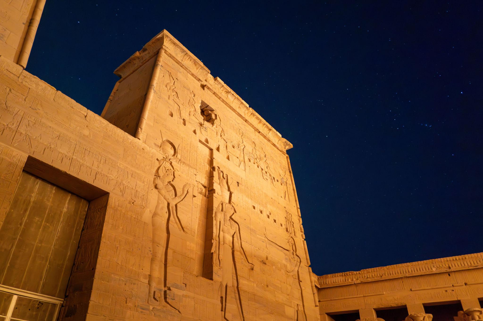 Illuminated Philae Temple pylon with carved Egyptian deity reliefs under a starry night sky