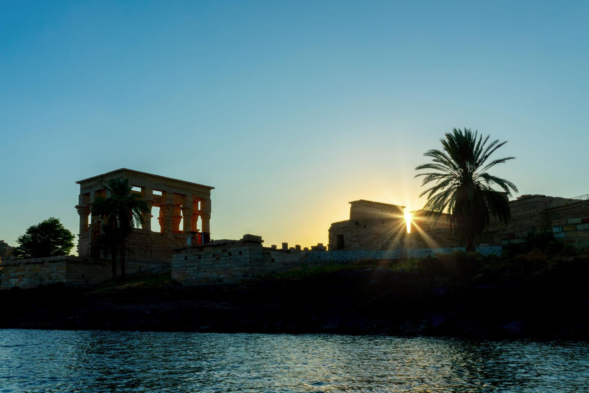 Philae Temple's Kiosk of Trajan silhouetted at sunset beside the Nile with palm trees