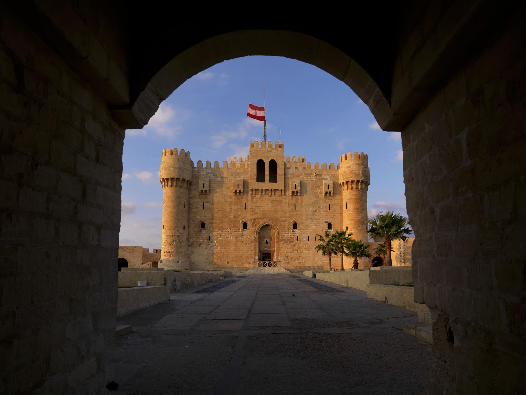 Qaitbay Citadel's main facade framed by a stone archway at dusk with flag flying