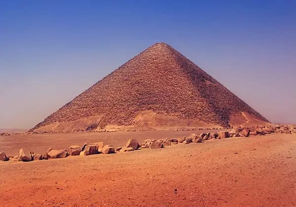 The Red Pyramid of Dahshur rises sharply against a vivid blue sky over the sandy desert.