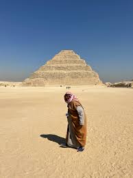 A person in traditional Egyptian attire stands before the Step Pyramid of Saqqara in the desert.
