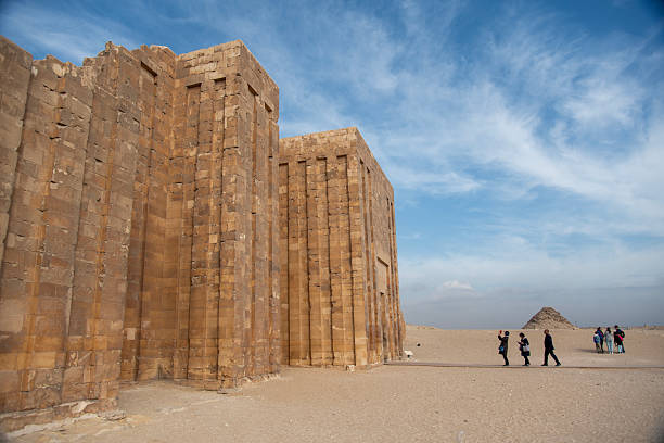 Tourists walk past the towering ancient stone enclosure wall at Saqqara with a pyramid beyond