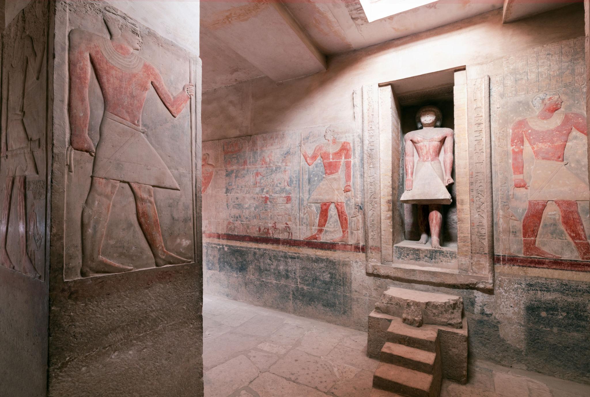Ancient Egyptian painted reliefs and a standing statue inside a dimly lit tomb chamber