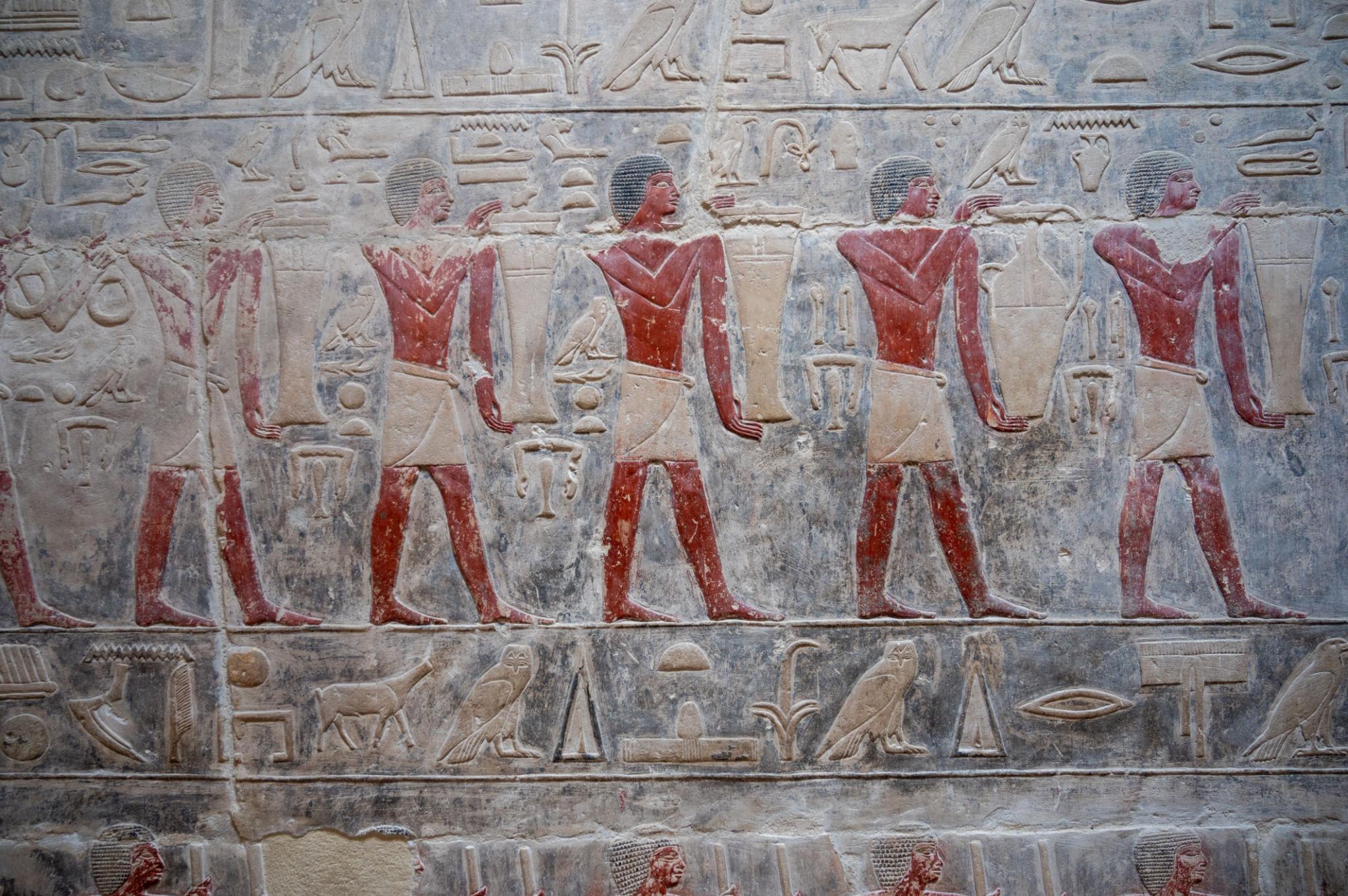 Ancient Egyptian painted relief showing four red-pigmented figures carrying tall vessels amid hieroglyphs
