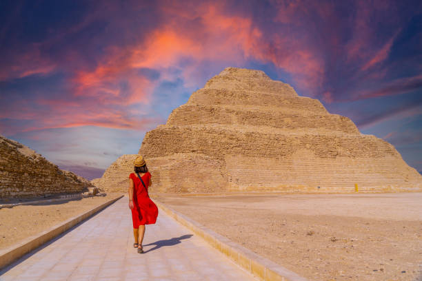 A woman in a red dress walks toward the Step Pyramid of Saqqara under a vivid sunset sky