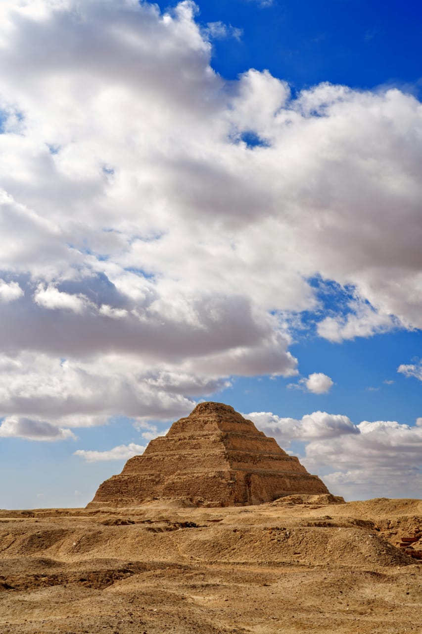 The Step Pyramid of Saqqara rises from sandy desert terrain beneath a dramatic cloudy blue sky.
