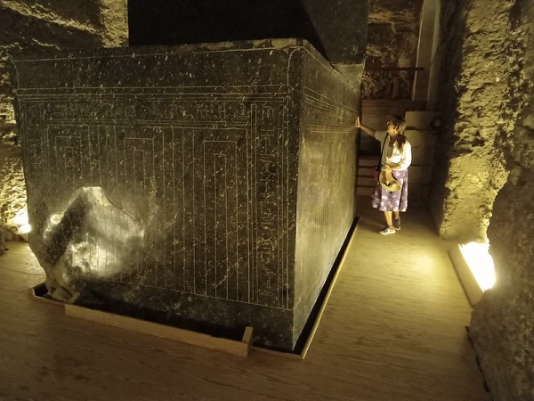 A visitor stands beside an enormous ancient granite sarcophagus in a dimly lit underground chamber.