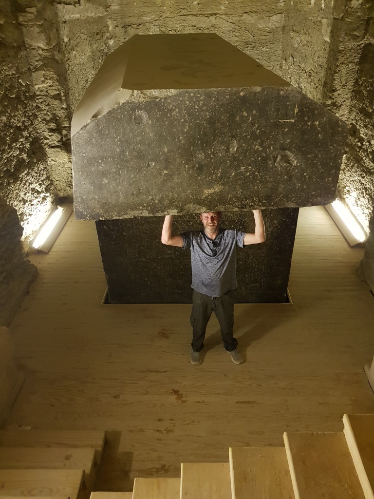 A smiling man poses as if lifting a massive ancient granite sarcophagus lid underground.
