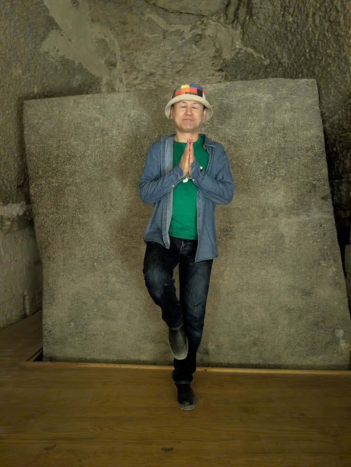 A visitor in a bucket hat holds hands in prayer pose beside a massive granite sarcophagus block